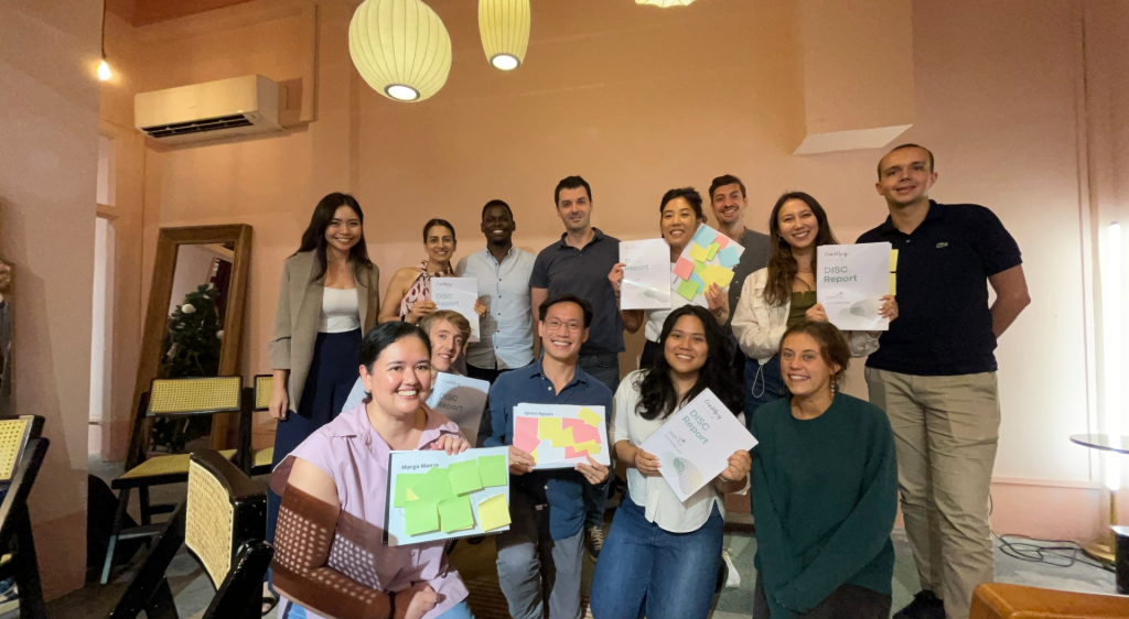 Group of diverse professionals smiling and holding DISC report documents during a corporate coaching session in a modern room. Keywords: corporate coaching, leadership development, team growth, DISC report.